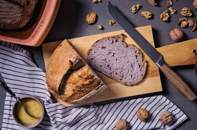 Frisch gebackenes Brot mit Walnüssen auf Holzbrett, daneben Honig und gestreiftes Tuch auf grauem Untergrund.