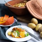 Keramiktopf mit Kartoffelauflauf, serviert auf einem weißen Teller mit Kürbis und frischem Gemüse, daneben Kartoffeln.