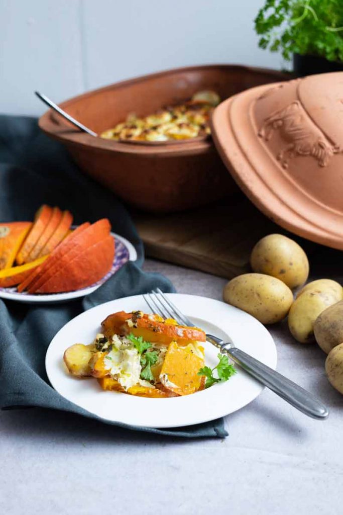 Keramiktopf mit Kartoffelauflauf, serviert auf einem weißen Teller mit Kürbis und frischem Gemüse, daneben Kartoffeln.