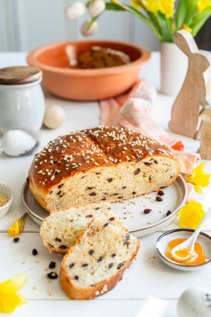Rosinenbrot auf einem Teller, dekoriert mit Hagelzucker, umgeben von Frühlingsdekorationen wie Narzissen und einem Keramikei.