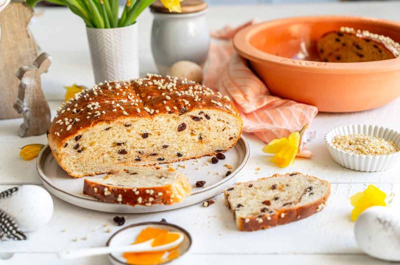 Osterbrot mit Rosinen und Hagelzucker, angeschnitten auf einem Teller, umgeben von Ostereiern und Narzissen.