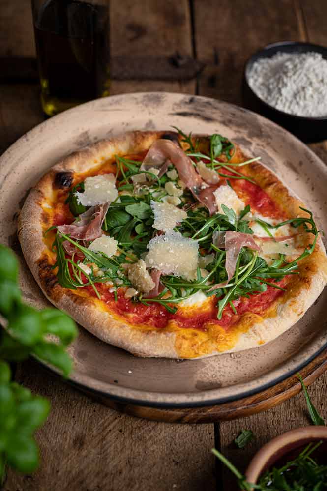 Pizza mit Rucola, Prosciutto und Parmesan, auf einem Teller serviert, daneben Olivenöl und Mehl in Schalen.