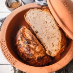 Frisch gebackenes Brot in einem Römertopf, daneben Gewürze und Mehl in Schälchen auf einem weißen Holztisch.