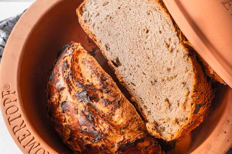 Frisch gebackenes Brot in einem Römertopf, daneben Gewürze und Mehl in Schälchen auf einem weißen Holztisch.