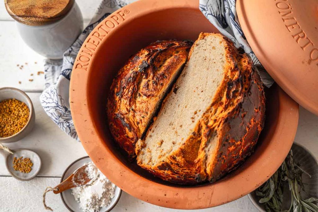 Brotlaib im Römertopf mit einer Scheibe geschnitten, umgeben von Mehl und Gewürzen auf weißem Holztisch.