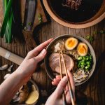 Person isst Ramen mit Stäbchen; Nudeln, Ei, Schweinefleisch, Frühlingszwiebeln in Schüssel. Knoblauch und Messer daneben.