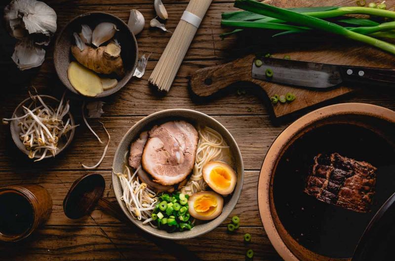 Schüssel mit Ramen, Schweinefleisch, Ei, Lauchzwiebeln und Sprossen, umgeben von Zutaten wie Knoblauch und Ingwer.