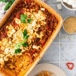 Auflaufform mit überbackenem Gericht aus Hackfleisch, Tomaten, Feta und Kräutern auf Tischdecke; ein Teller serviert daneben.
