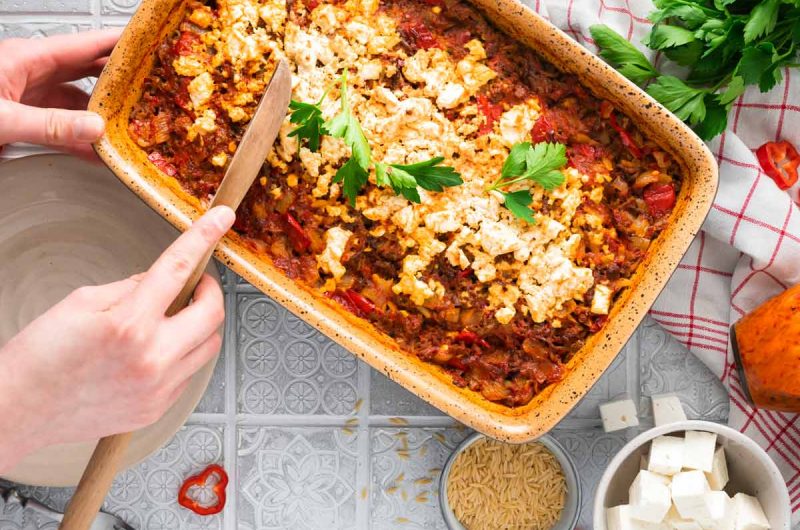 Eine Person schneidet einen Auflauf mit Tomaten, Paprika und Feta in einer Backform an, daneben frische Petersilie.