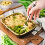 Hand mit Gabel in einem gebackenen grünen Auflauf mit Käsekruste, auf einem hölzernen Brett serviert.
