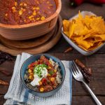 Schüssel mit Chili con Carne, serviert auf Reis mit Sauerrahm. Im Hintergrund Tortilla-Chips und Paprika.