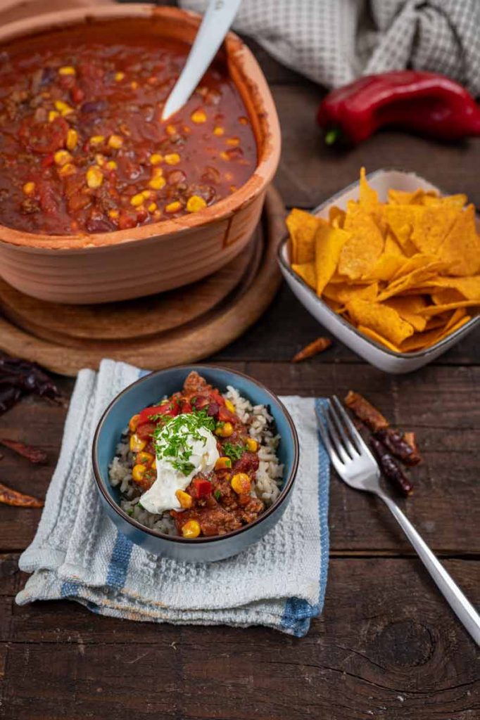 Schüssel mit Chili con Carne, serviert auf Reis mit Sauerrahm. Im Hintergrund Tortilla-Chips und Paprika.