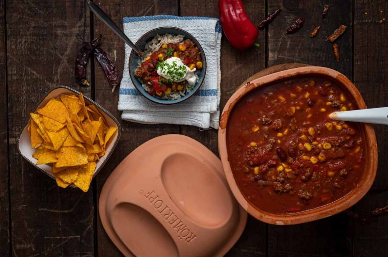 Schüssel mit Chili con Carne neben Tortillachips und einer Servierschale auf Holzuntergrund.