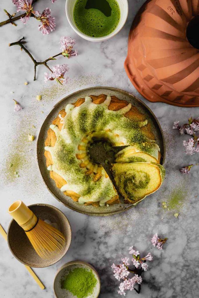 Gugelhupf mit grünem Matcha-Pulver, dekoriert mit Zuckerguss. Daneben Blütenzweige und eine Schale Matcha-Tee.