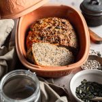 Vollkornbrot in einem Tontopf, umgeben von Kürbiskernen, Sonnenblumenkernen und einem Glas mit Leinsamen.