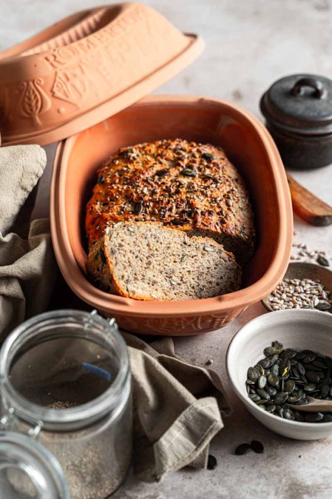 Vollkornbrot in einem Tontopf, umgeben von Kürbiskernen, Sonnenblumenkernen und einem Glas mit Leinsamen.