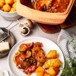 Römertopf mit Gemüseauflauf, serviert auf einem Teller mit Kroketten und Käse, umgeben von Zutaten wie Pilzen und Paprika.