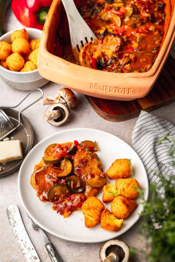 Römertopf mit Gemüseauflauf, serviert auf einem Teller mit Kroketten und Käse, umgeben von Zutaten wie Pilzen und Paprika.