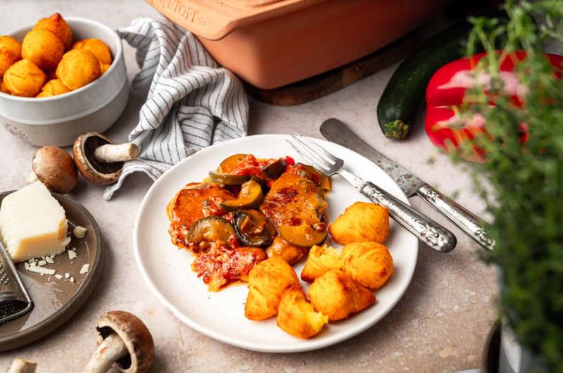 Teller mit Gemüseauflauf und Kroketten, umgeben von Pilzen, Käse, roter Paprika und frischen Kräutern.
