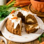 Möhrchenkuchen mit Frischkäse-Füllung und Zuckerguss, dekoriert mit Marzipanmöhren, auf Holzplatte, Karotten im Hintergrund.