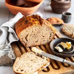 Frisch gebackenes Brot auf einem Holzbrett, mit Butter bestrichen. Im Hintergrund ein Tontopf und Gewürze.