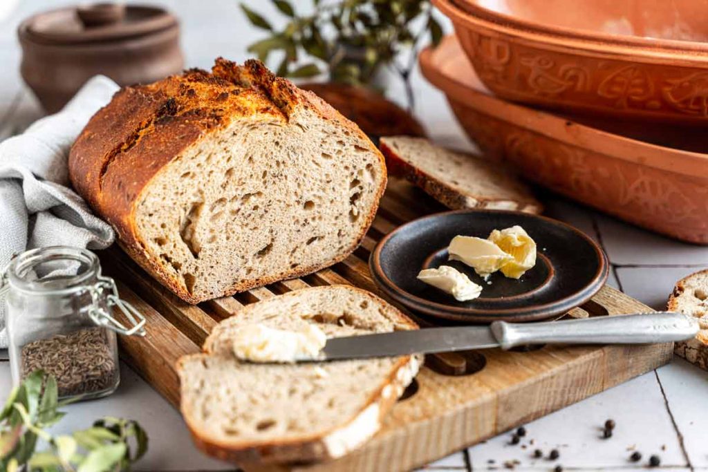 Frisches, geschnittenes Brot auf einem Holzbrett, daneben ein Teller mit Butter und ein Messer.