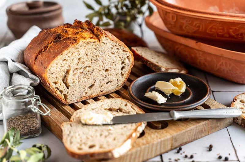 Frisches, geschnittenes Brot auf einem Holzbrett, daneben ein Teller mit Butter und ein Messer.