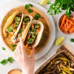 Hand greift nach einem von drei Tacos auf Holzplatte, umgeben von Zutaten wie Limetten und Karottenstreifen.