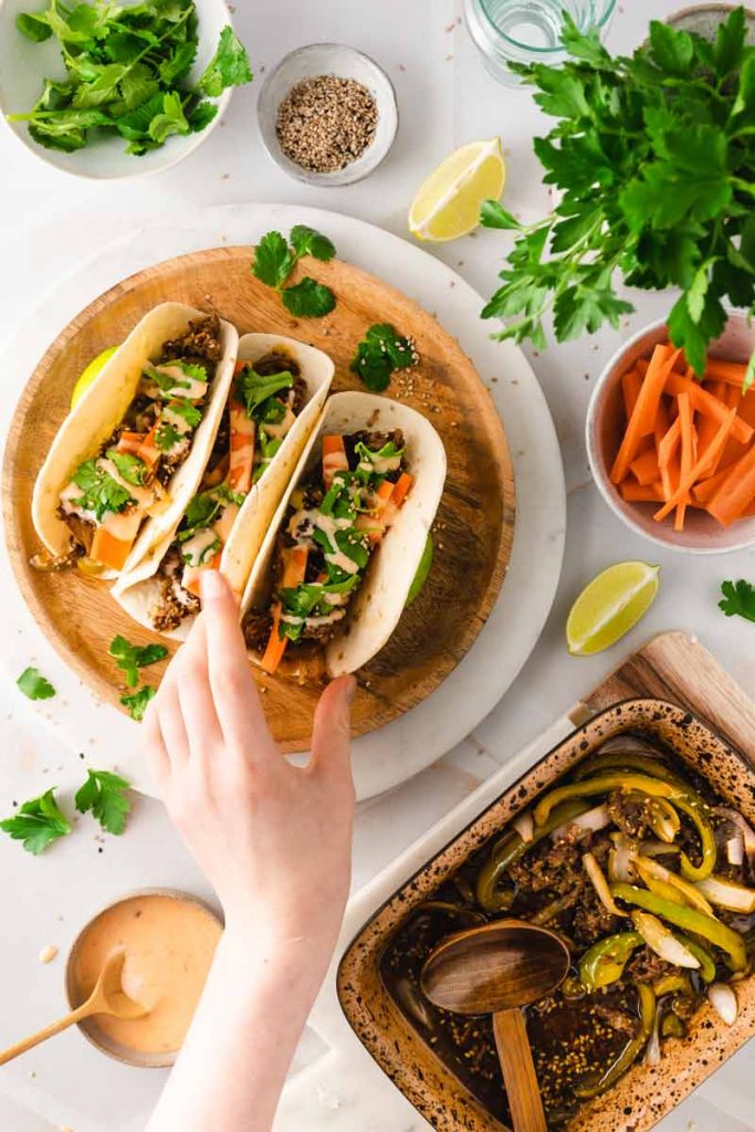 Hand greift nach einem von drei Tacos auf Holzplatte, umgeben von Zutaten wie Limetten und Karottenstreifen.