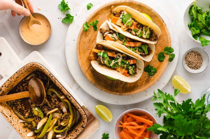 Drei Tacos mit Gemüse und Koriander auf Holzbrett, umgeben von Soße, Limetten und frischen Zutaten.