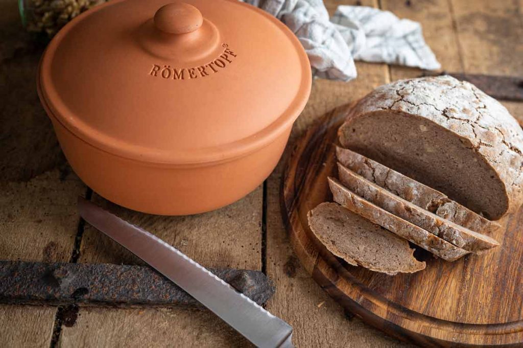 Ein Laib Brot, teilweise geschnitten, auf einem Holzbrett neben einem Römertopf und einem Brotmesser.