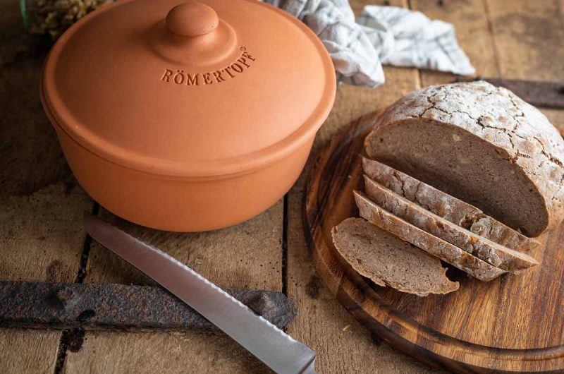 Ein Laib Brot, teilweise geschnitten, auf einem Holzbrett neben einem Römertopf und einem Brotmesser.