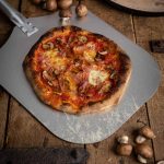 Pizza mit Pilzen und Schinken auf einem Pizzablech, daneben frische Champignons auf rustikalem Holztisch.