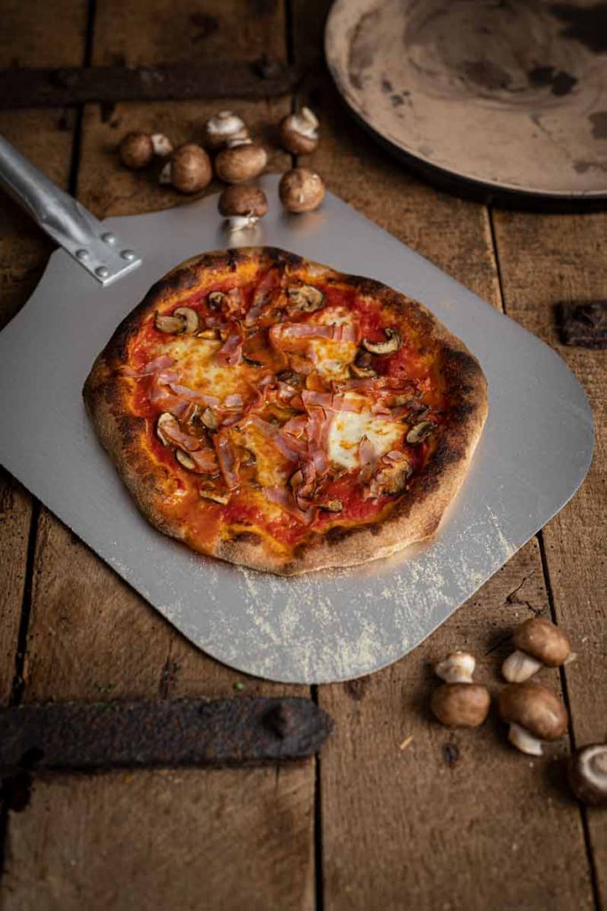 Pizza mit Pilzen und Schinken auf einem Pizzablech, daneben frische Champignons auf rustikalem Holztisch.