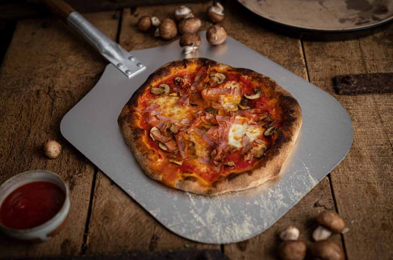 Holzpizza mit Schinken, Käse und Pilzen auf Pizzaschieber, daneben Tomatensauce und Pilze auf Holztisch.