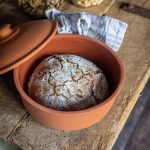 Rundes Brot in einem offenen Tontopf auf einem Holztisch, mit Nussgläsern im Hintergrund.