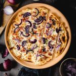 Flammkuchen mit roten Zwiebeln und Speck auf Teller, umgeben von Zutaten wie Zwiebeln und Gewürzen.
