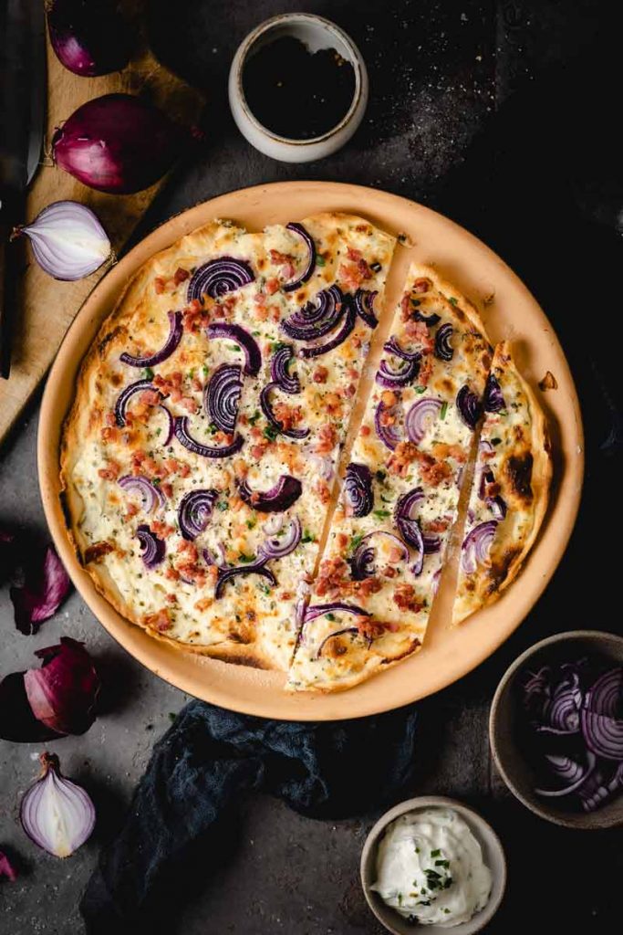 Flammkuchen mit roten Zwiebeln und Speck auf Teller, umgeben von Zutaten wie Zwiebeln und Gewürzen.