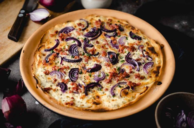 Flammkuchen mit roten Zwiebeln und Speck auf einem runden, braunen Teller, auf Holztisch angerichtet.