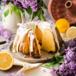 Kuchen mit Zitronenglasur auf Teller, umgeben von Fliederblüten und Zitronen. Hintergrund: Fliederstrauß und Gugelhupfform.