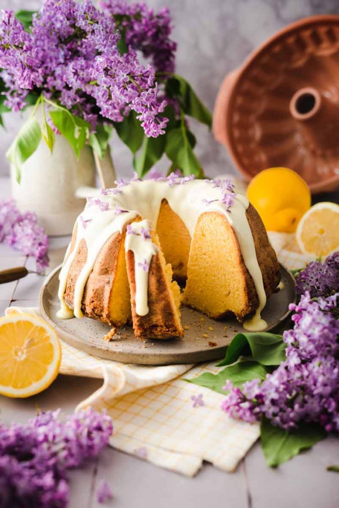 Kuchen mit Zitronenglasur auf Teller, umgeben von Fliederblüten und Zitronen. Hintergrund: Fliederstrauß und Gugelhupfform.