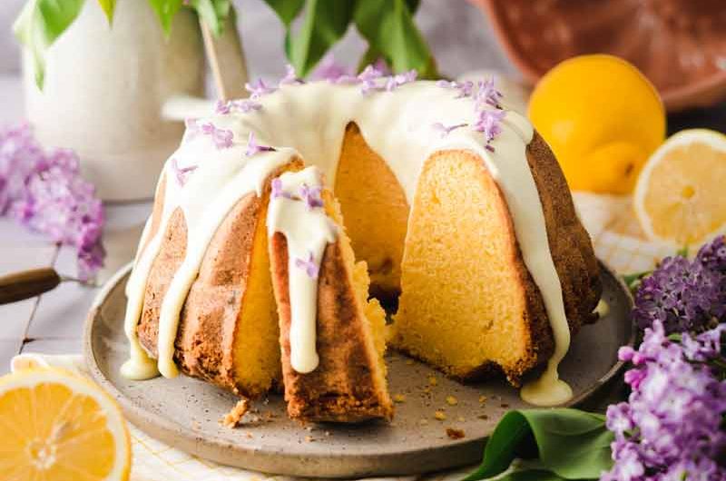 Kuchen mit Zitronenglasur auf Teller, umgeben von Fliederblüten und Zitronen. Hintergrund: Fliederstrauß und Gugelhupfform.