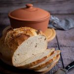 Laib Brot mit einigen Scheiben auf Holzbrett, im Hintergrund ein Tontopf, auf rustikalem Tisch.