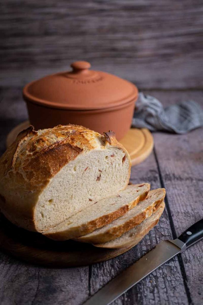 Laib Brot mit einigen Scheiben auf Holzbrett, im Hintergrund ein Tontopf, auf rustikalem Tisch.
