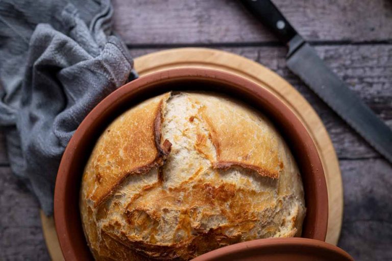 Frisch gebackenes Brot in einem Tontopf, daneben ein graues Tuch und ein Brotmesser auf einem Holztisch.