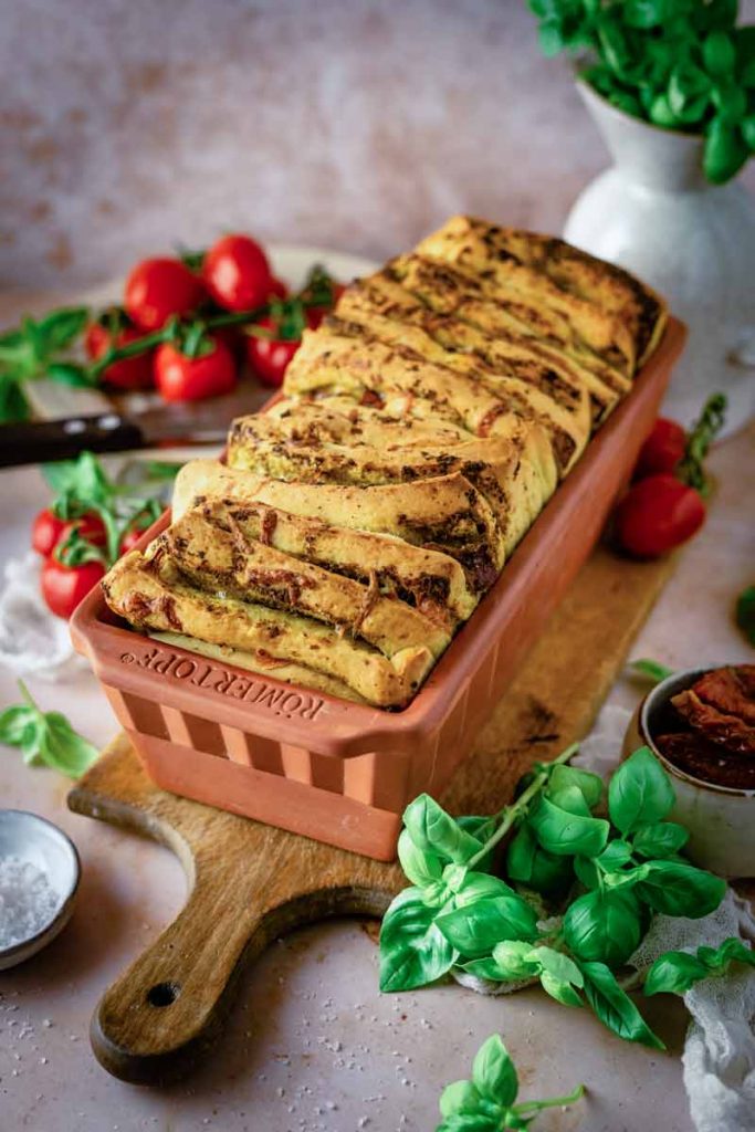 Faltbrot im Terrakotta-Backform, umgeben von Tomaten und Basilikum auf einem Holzbrett.