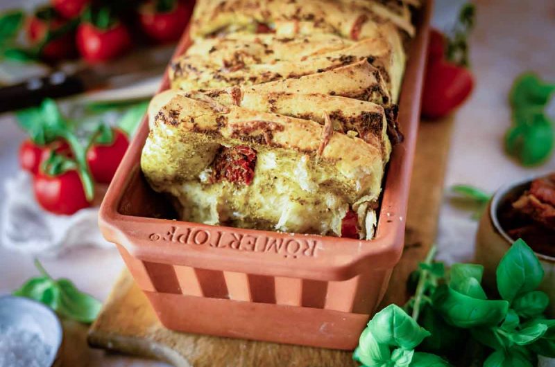 Herzhaftes Brot mit Tomaten und Kräutern in einer Terrakottaform, umgeben von frischem Basilikum und Tomaten.