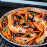 Gegrillte Hähnchenschenkel mit Karotten, Sesam und Kräutern in einer Pfanne auf einem Grill.