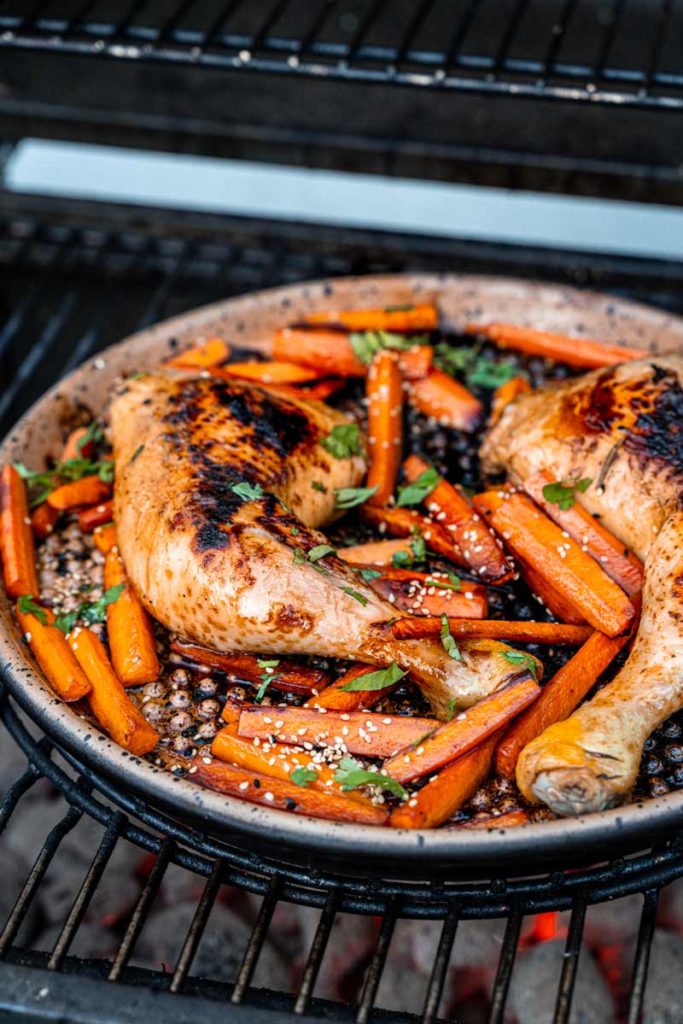 Gegrillte Hähnchenschenkel mit Karotten, Sesam und Kräutern in einer Pfanne auf einem Grill.