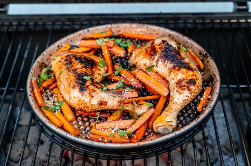 Gegrillte Hähnchenschenkel mit Karotten, garniert mit Sesam und Kräutern in einer runden Pfanne auf einem Grill.